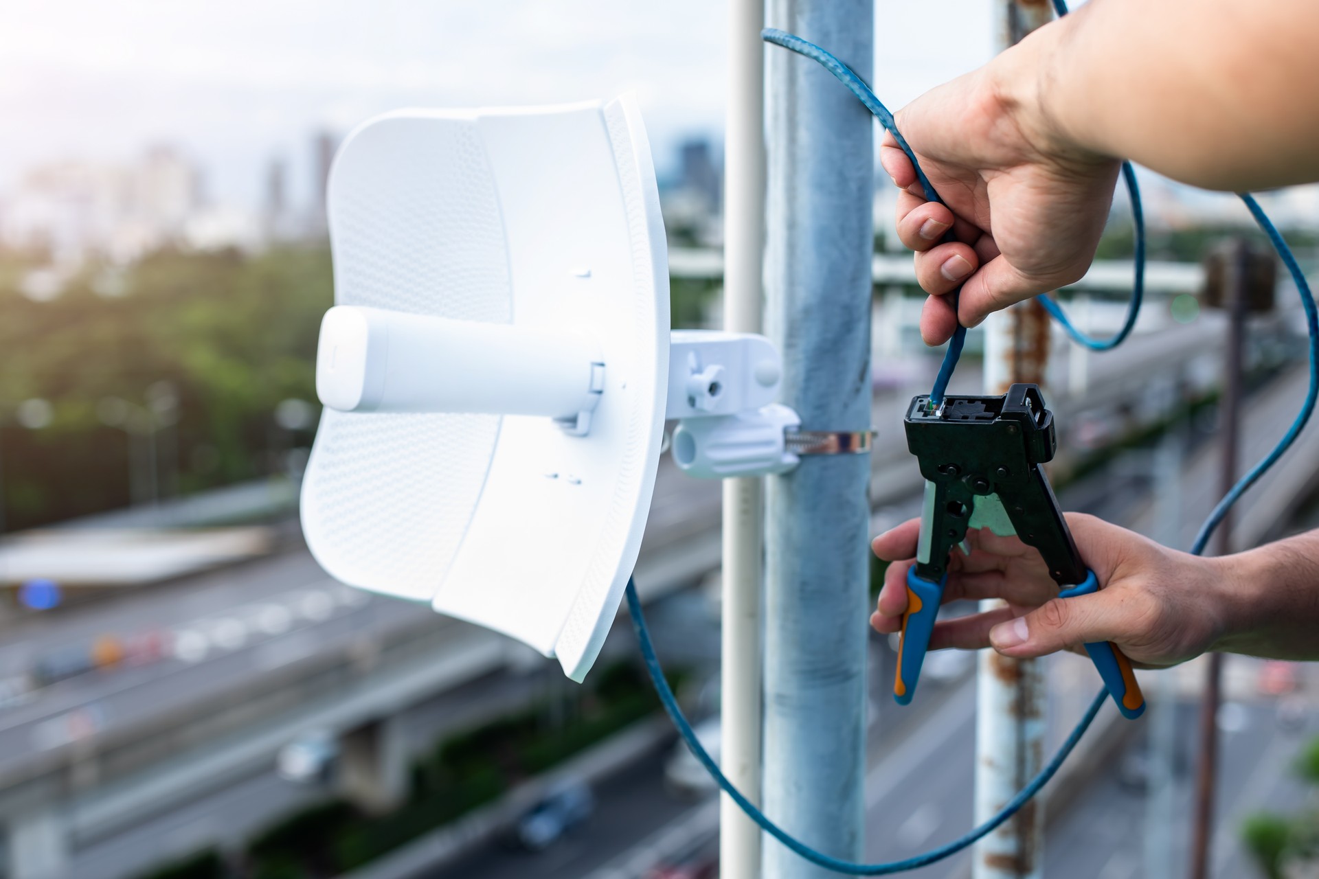 Selective focus to engineer's hand crimping RJ45 LAN cable with blurry WIFI transmitter dish to installation system. Connection procedure UTP cable.